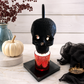 A black skull-shaped candle warmer lamp with glowing amber eyes and a red glittery candle base, displayed on a black square pedestal on a marble surface with seasonal decor including a small pumpkin, black fabric, and a vase in the background.