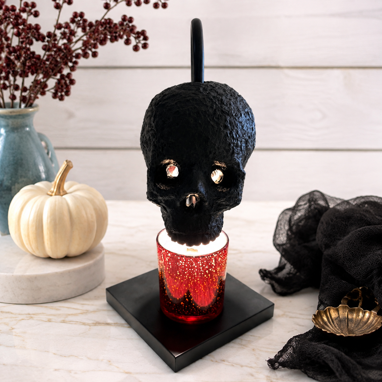 A black skull-shaped candle warmer lamp with glowing amber eyes and a red glittery candle base, displayed on a black square pedestal on a marble surface with seasonal decor including a small pumpkin, black fabric, and a vase in the background.