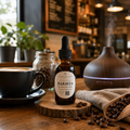 A small amber glass dropper bottle labeled 'BARISTA' stands on a wooden slice on a cafe table, surrounded by coffee beans, a black coffee cup, a diffuser, and a cloth with more coffee beans.