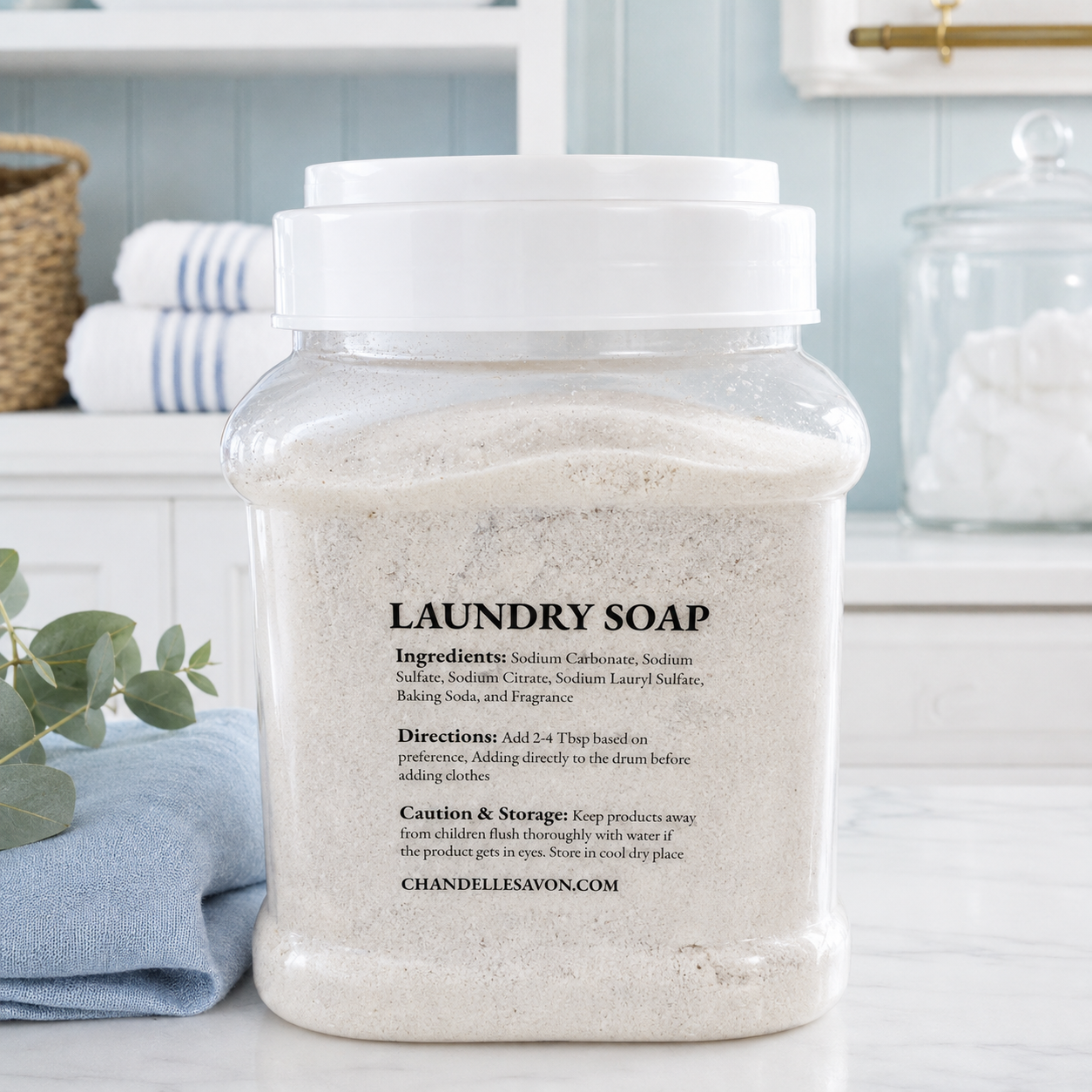 A clear plastic jar of Chandelle & Savon laundry soap with a white screw-top lid, filled with a light beige powder. The jar is placed on a white marble countertop in a bright, modern laundry room setting with a blue wall and folded towels in the background.
