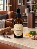 An amber glass dropper bottle labeled 'Chandelle & Savon Teak Mahogany' sits on a stone countertop in a warm, rustic living room setting with wood furniture, a leather chair, and natural ingredients like lavender, citrus peel, and ginger.