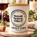 A white ceramic candle jar with a light wood lid, featuring a black printed circular logo for Highland Reserves Winery and the text 'SWEET INKS'. The candle is styled on a marble coaster with a cork, a glass of red wine, and grapes in a blurred background.