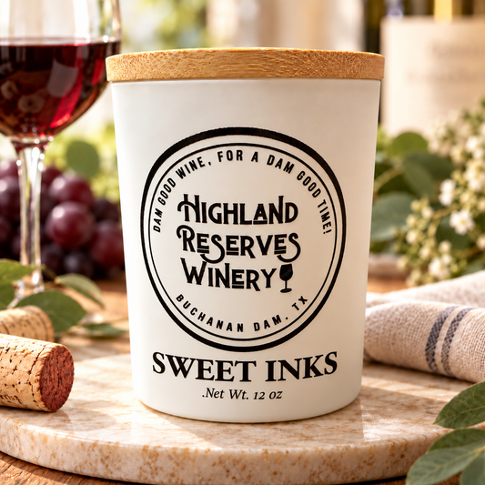 A white ceramic candle jar with a light wood lid, featuring a black printed circular logo for Highland Reserves Winery and the text 'SWEET INKS'. The candle is styled on a marble coaster with a cork, a glass of red wine, and grapes in a blurred background.